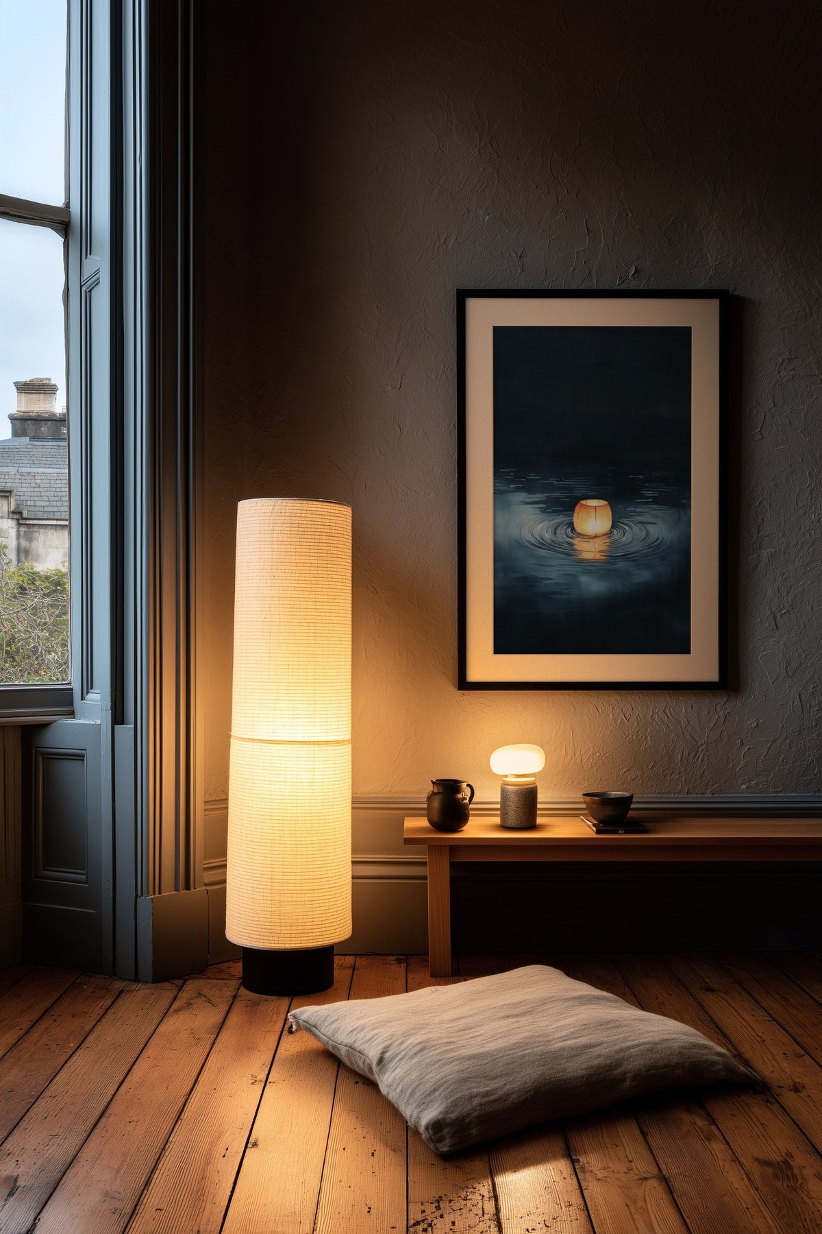 Dark Japandi corner at dusk with a tall glowing rice paper floor lantern beside a framed indigo ink print of a single floating lantern