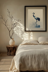 Indigo Crane print in a black frame above a bed with linen bedding and a vase of bare branches, Yugen Print