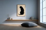 A framed art print of a black cat silhouette on a beige background, hanging on a light blue-gray wall in a minimalist room with a wooden floating shelf, books, and a floor cushion.