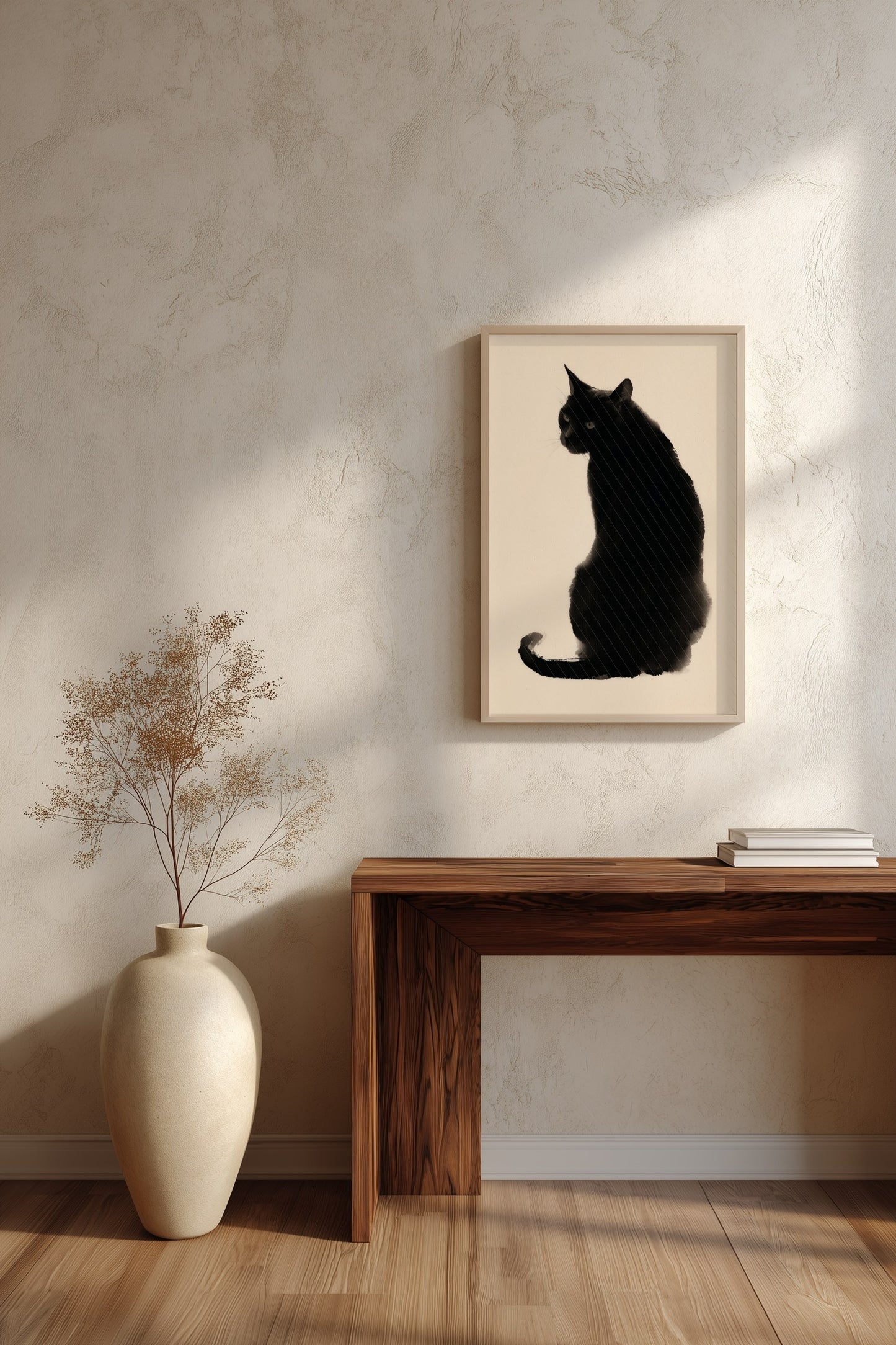 A framed black cat silhouette art print hangs on a textured beige wall above a wooden console table, with a cream vase of dried branches on the left and a stack of books on the right.
