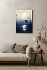 Sumi-e style indigo ink print of a crane silhouetted beneath a full moon, black frame above a pale linen sofa with a wooden side table and ceramic lamp, Yugen Print