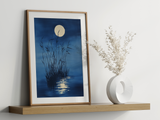 Sumi-e style indigo ink print of tall reeds beneath a full moon, oak frame leaning on a floating wood shelf beside a white ceramic vase, Yugen Print