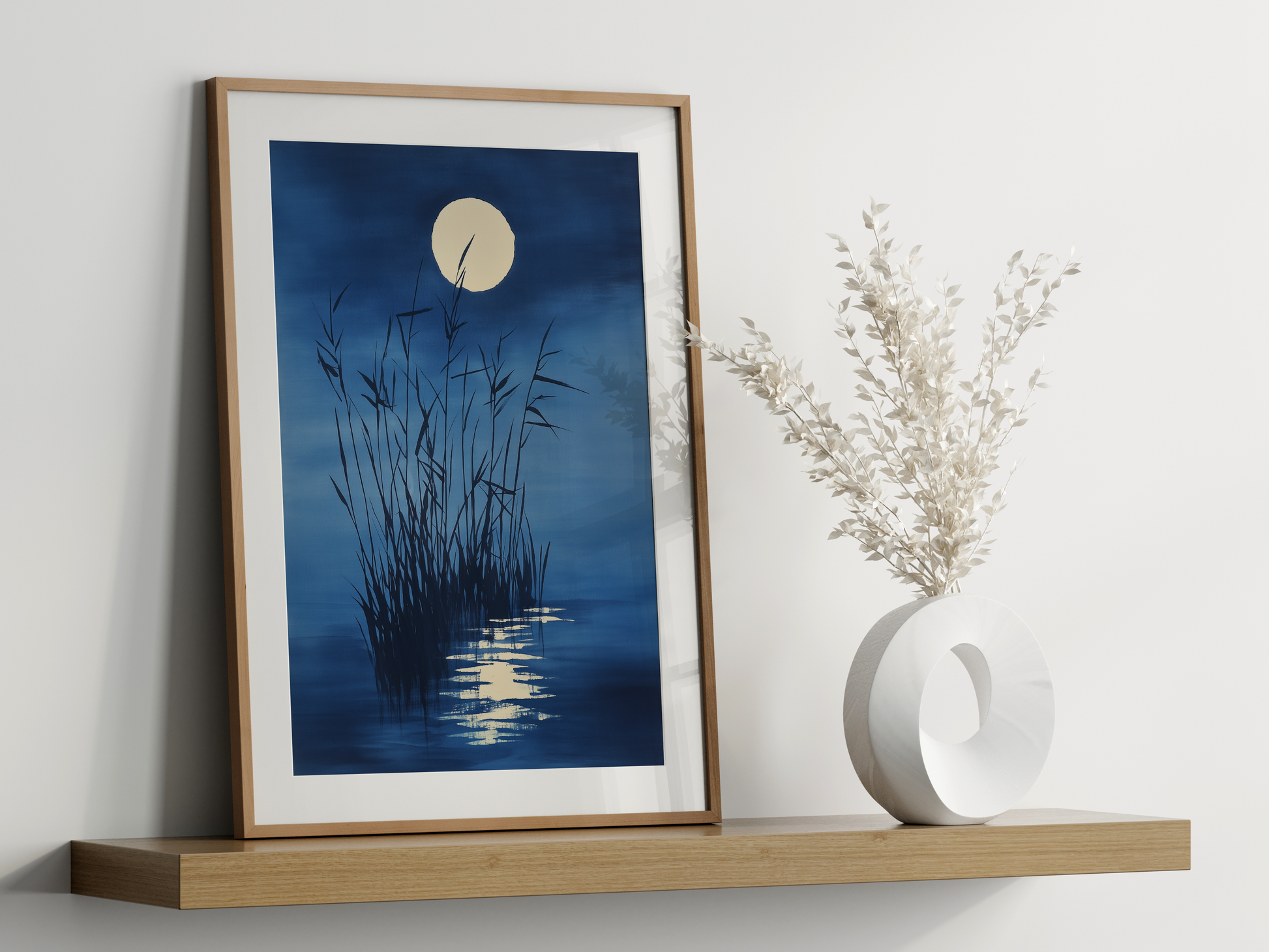 Sumi-e style indigo ink print of tall reeds beneath a full moon, oak frame leaning on a floating wood shelf beside a white ceramic vase, Yugen Print