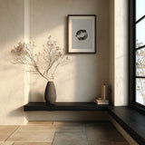 A dark wood framed black and white fugu ensō ink art print hangs on a warm ivory wall above a black floating shelf with a stone vase of dried branches beside a window in a japandi entryway nook.