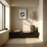 A framed black and white fugu ensō ink art print hangs on a warm ivory wall above a low black shelf with a folded cushion and books, beside a bare branching tree in a minimalist reading nook.