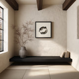 A framed black and white fugu ensō ink art print hangs on a textured cream wall beneath dark wood ceiling beams, above a black bench with a vase of branches in a quiet meditation nook.