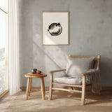 A framed black and white fugu ensō ink art print hangs on a soft grey-beige wall above a light wood armchair and a small side table with a tea set in a sunlit reading corner.