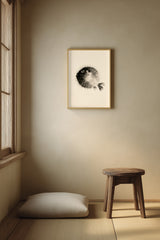A framed black and white pufferfish art print hangs on a beige wall in a minimalist Japanese-style room with a wooden stool and a floor cushion.