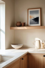 The Narrow Crossing sumi-e print framed on the wall above a wood shelf in a warm ivory kitchen corner